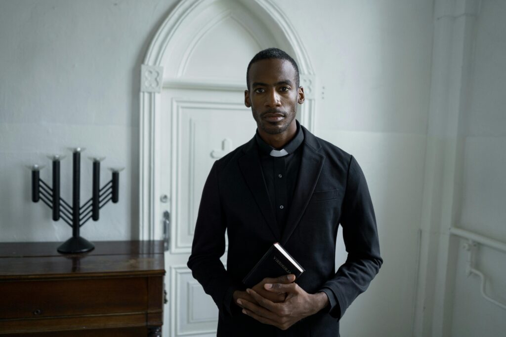 A solemn priest in black attire holds a Bible, standing inside a church.