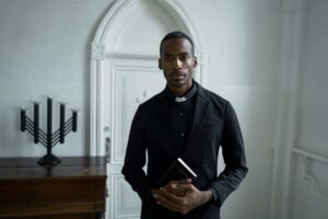 A solemn priest in black attire holds a Bible, standing inside a church.